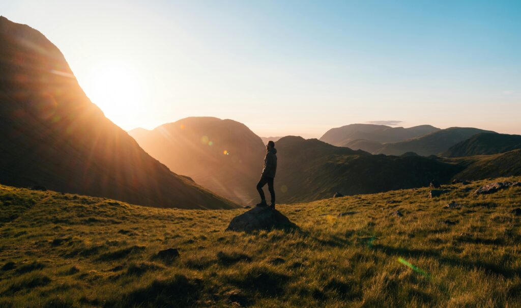 Silhouette Photography of Person Standing on Green Grass in Front of Mountains during Golden Hour